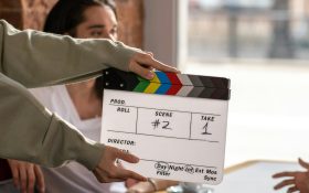 a clapperboard is centre of frame, behind are two people at a cafe table next to a window. One has only their hand seen, the other is of indeterminate gender and race. conscious casting.