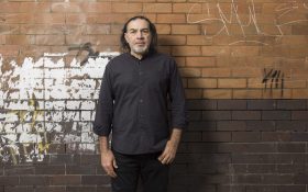 Khaled Sabsabi, a middle-aged Lebanese Australian man with thin, should-length black hair, gray beard, wearing black shirt and pants, standing against a graffitied brick wall.