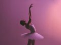 Ballerina poses,. leaning to the right with an arm raised. Pink background.