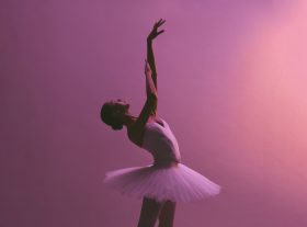 Ballerina poses,. leaning to the right with an arm raised. Pink background.