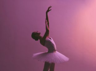 Ballerina poses,. leaning to the right with an arm raised. Pink background.