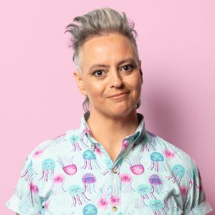 against a pink background a woman with a grey mohawk and a short sleeved colourful shirt smiles at the camera. Geraldine Hickey