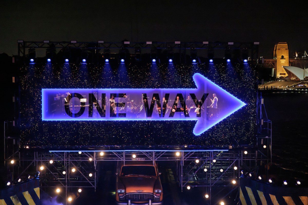 An orchestra playing behind a huge purple see-through arrow saying 'one way' and point to the right above an outdoor stage with a New York yellow cab in the middle. In the background to the right we can see part of the Sydney Harbour Bridge. Guys & Dolls.