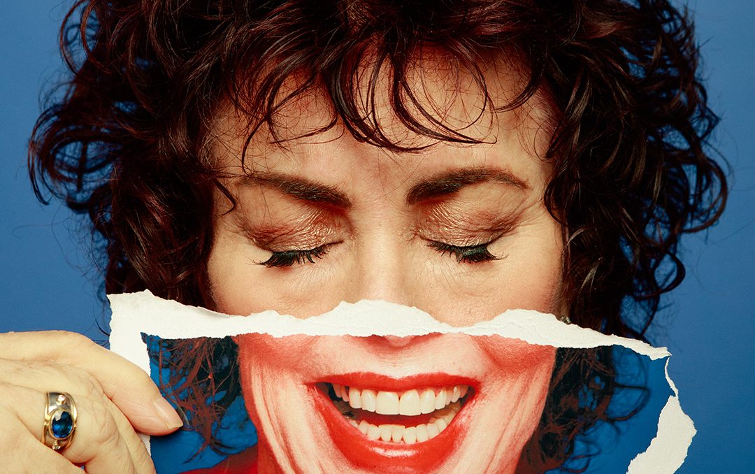 A Caucasian woman with short curly dark hair has her eyes closed and is holding a picture over the lower half of her face with a big smiling mouth on it. Ruby Wax