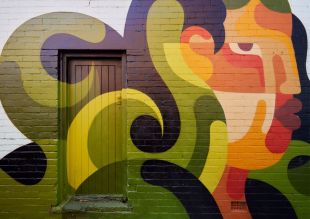 Benalla Street Art Festival 2025 will unveil 10 new major pieces in locations across the regional Victorian city. Pictured is an artwork by REKA, unveiled in 2024; w stylised image of a human face in profile, with the hair painted in swirls of orange, while the facial features, including eyes, ears, nose and lips are painted in shades of red, white, brown and orange.