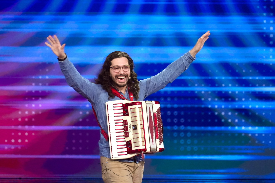 A man with glasses and curly brown hair wearing a blue shirt with his arms in the air. He is carrying an accordion.