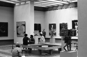 A black and white photograph, taken in 1962, of a large modern light-filled gallery space showing an exhibition with paintings on the walls and sculptures installed in the space. There are three well-dressed people in the gallery looking at the art.