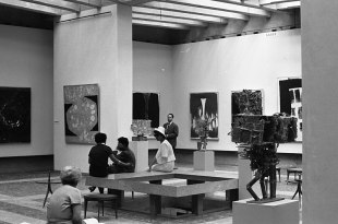 A black and white photograph, taken in 1962, of a large modern light-filled gallery space showing an exhibition with paintings on the walls and sculptures installed in the space. There are three well-dressed people in the gallery looking at the art.