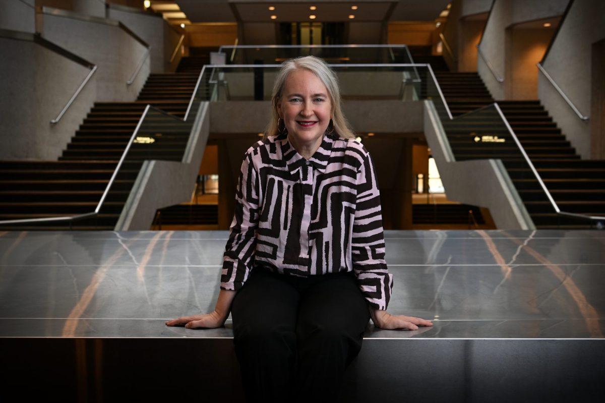 A woman in a black and white patterned shirt with grey shoulder length straight hair leans against a divider with some steps leading upwards behind her on both right and left. Rachel Healy.