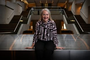 A woman in a black and white patterned shirt with grey shoulder length straight hair leans against a divider with some steps leading upwards behind her on both right and left. Rachel Healy.