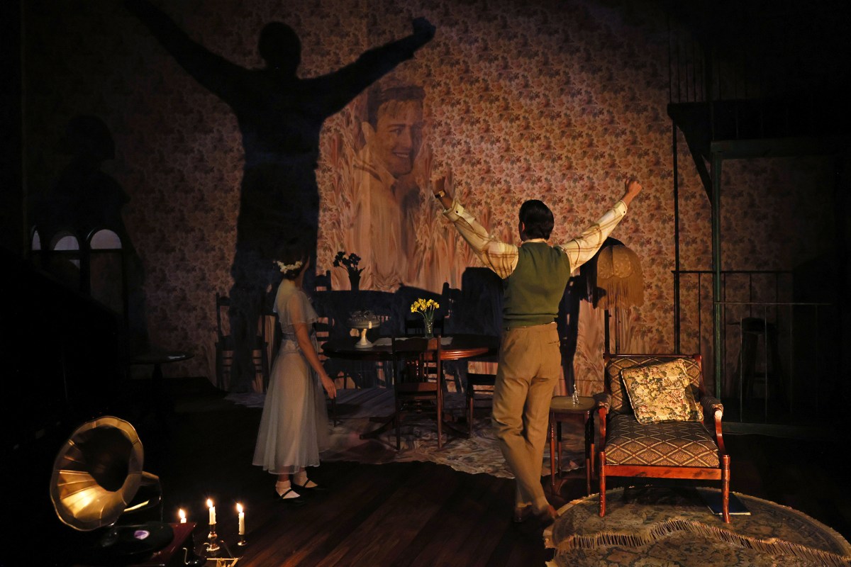 A scene from Ensemble Theatre’s 2025 production of 'The Glass Menagerie'. A young man stands with his back to the camera, arms upraised; his shadow falls dramatically on the wall behind him. A slender, waiflike young woman, her face turned away from the camera, stares at his dramatically lit shadow.