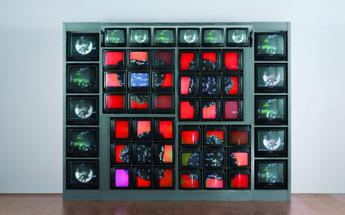 An installation artwork by Nam June Paik of 52 television screens stacked against a wall, each screen is displaying something different, but some are grouped together to form a coherent shape, such as a heart.