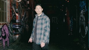 A young Asian man wearing a blue checked flannel shirt standing in a laneway with graffiti. Chris Nguyen