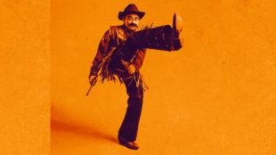 Against a yellow background, a woman dressed as a man dressed in a cowboy costume has one of his legs in the air, mid kick.
