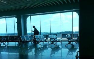 An airport waiting lounge with green hue and a lone figure looking at something in their hand in the distance. US travel.