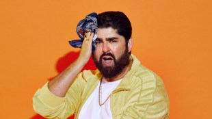 A beardy brunette in a white t-shirt and yellow shirt. He is holding a blue hankie to his forehead.