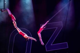 Three trapeze artists swinging in the air. They are wearing red leotards. In the background the word 'Oz" can be seen.