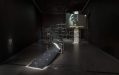A steel installation with a video projection screen on the right hand side, resembling a ramp for motorcycle stunts. It is installed inside a dark gallery space.