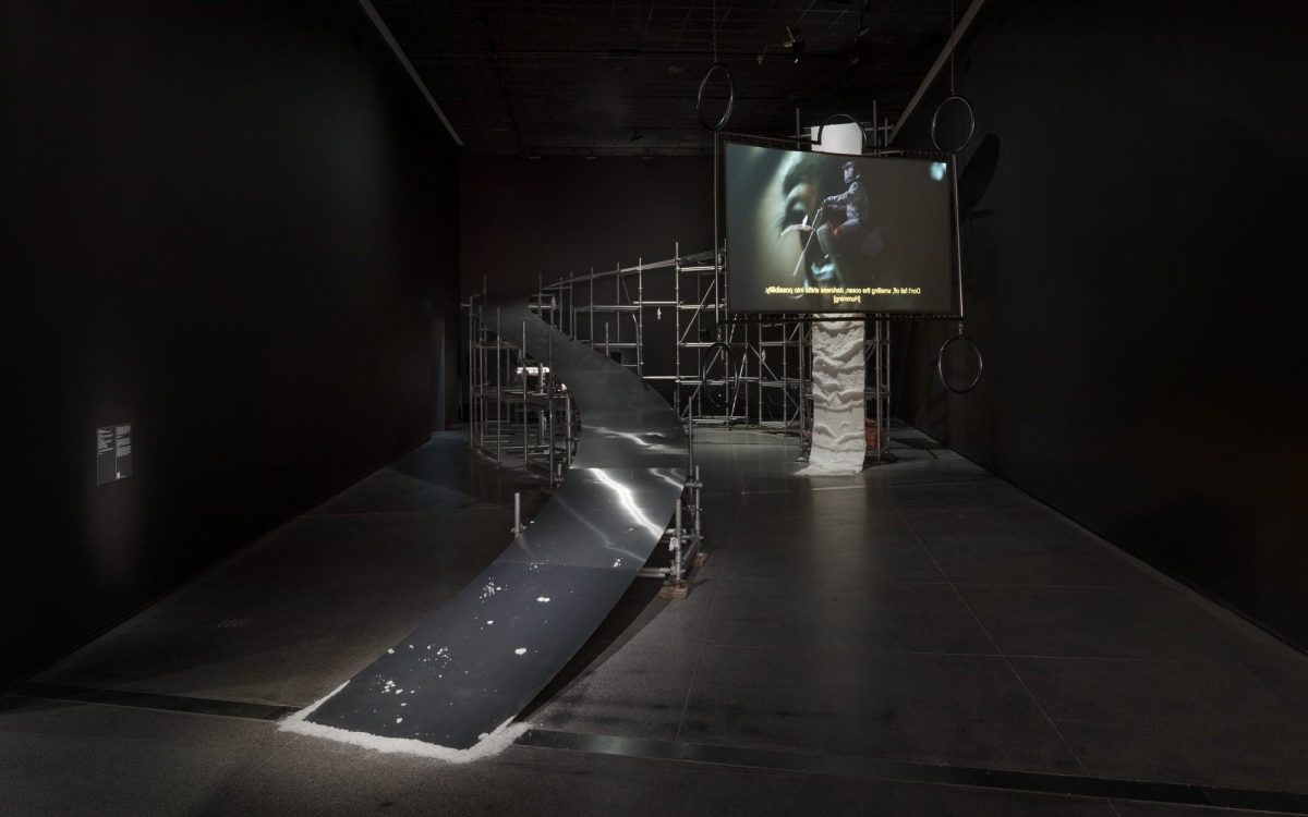 A steel installation with a video projection screen on the right hand side, resembling a ramp for motorcycle stunts. It is installed inside a dark gallery space.