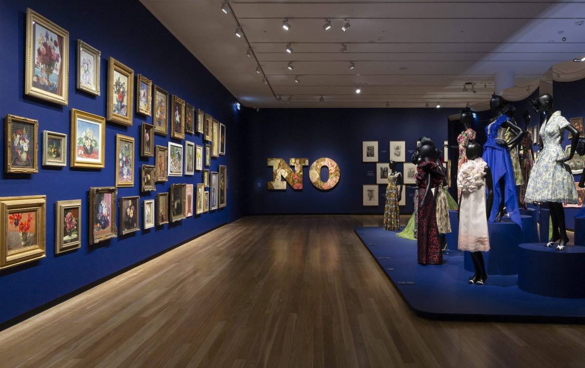 Installation shot of an exhibition with paintings and garments on mannequins on both sides. The walls are painted an ultramarine blue, and large text “NO” is installed on the back wall.