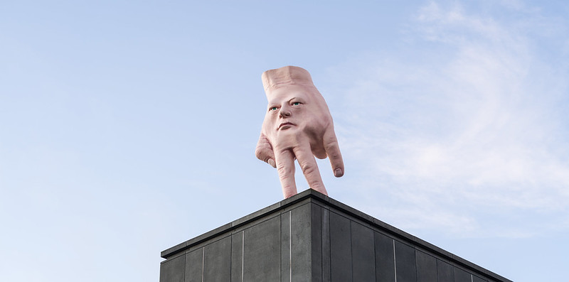 'Quasi' by Ronnie Van Hout, Dark Mofo 2025. A giant fusion of the artist's hand, his face embedded in its palm, stands on its fingertips at the edge of a city building.