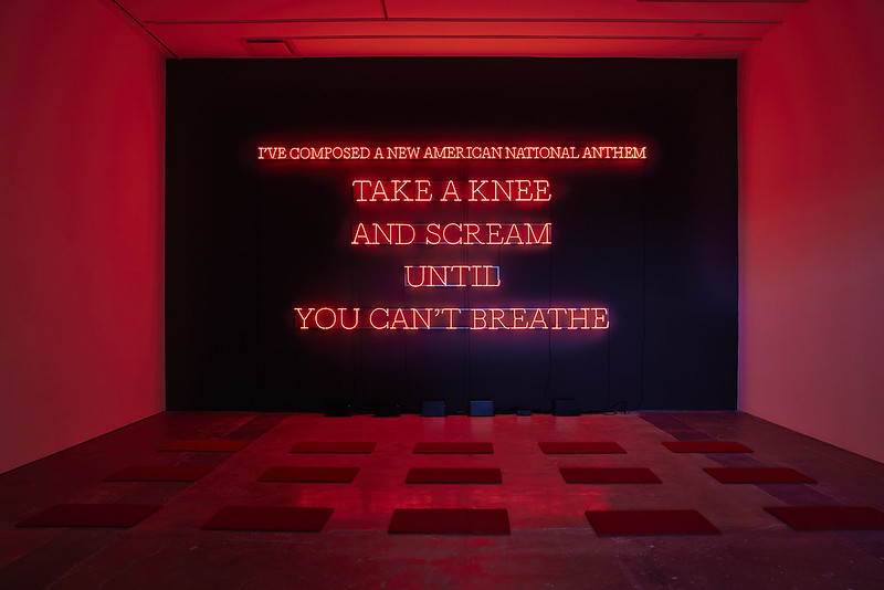 'Neon Anthem' by Nicholas Galanin, Dark Mofo 2025. Red text in capital letters constructed of glowing neon tubes spell out the words 'I've composed a new Americal national anthem. Take a knee and scream until you can't breathe.' A series of mats on the floor in front of the installation are visible, where audience members are encouraged to kneal and scream, thus creating the artwork in question.