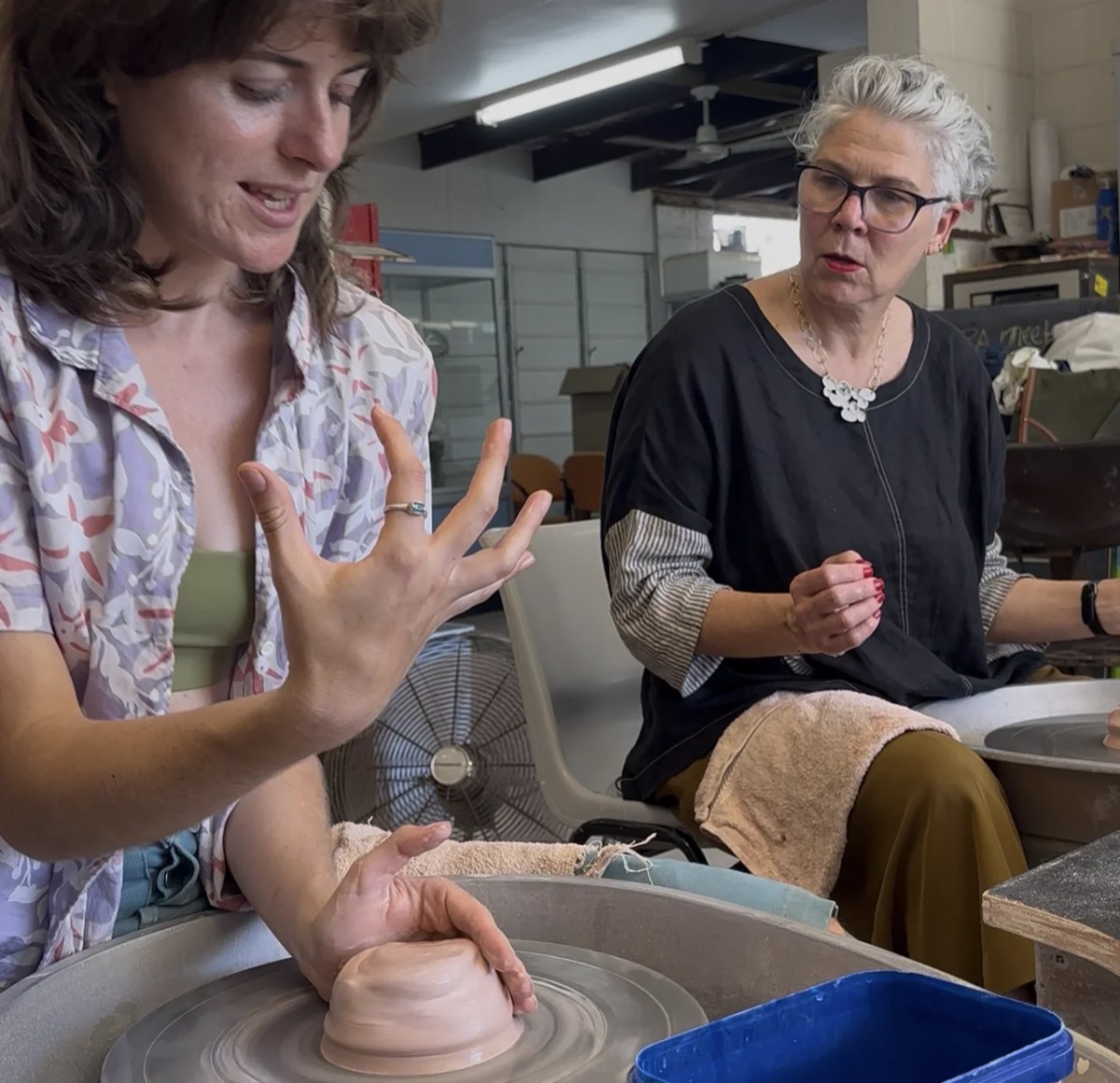 Young non-binary person teaching older woman how to create on a potters wheel. Baylee Griffin