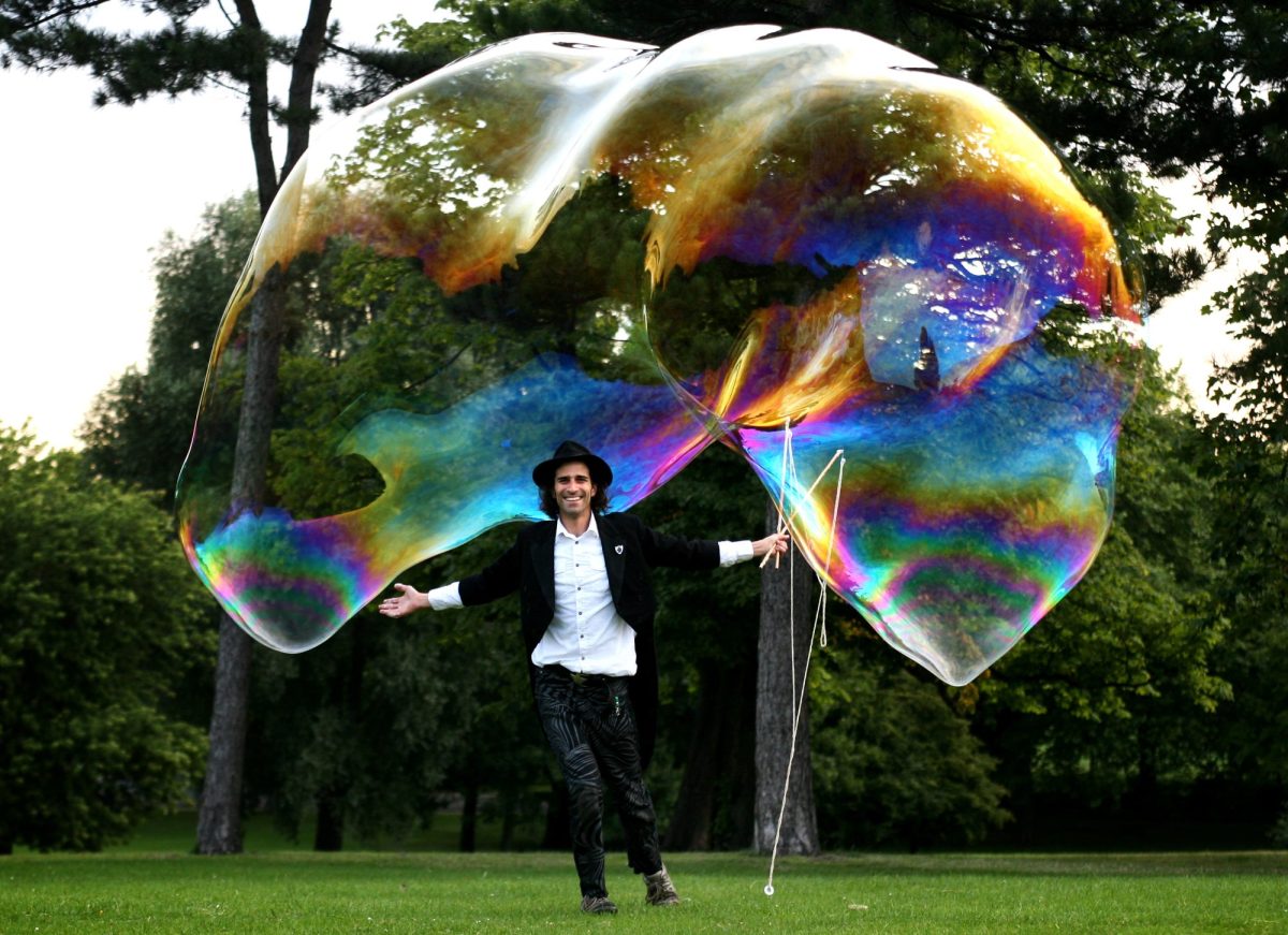A man in a white shirt and black jacket and pants runs through a park with a giant bubble around him.