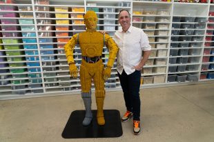 A life-size lego version of C3PO with its creator Ryan McNaught, standing next to it with a white top and black pants.