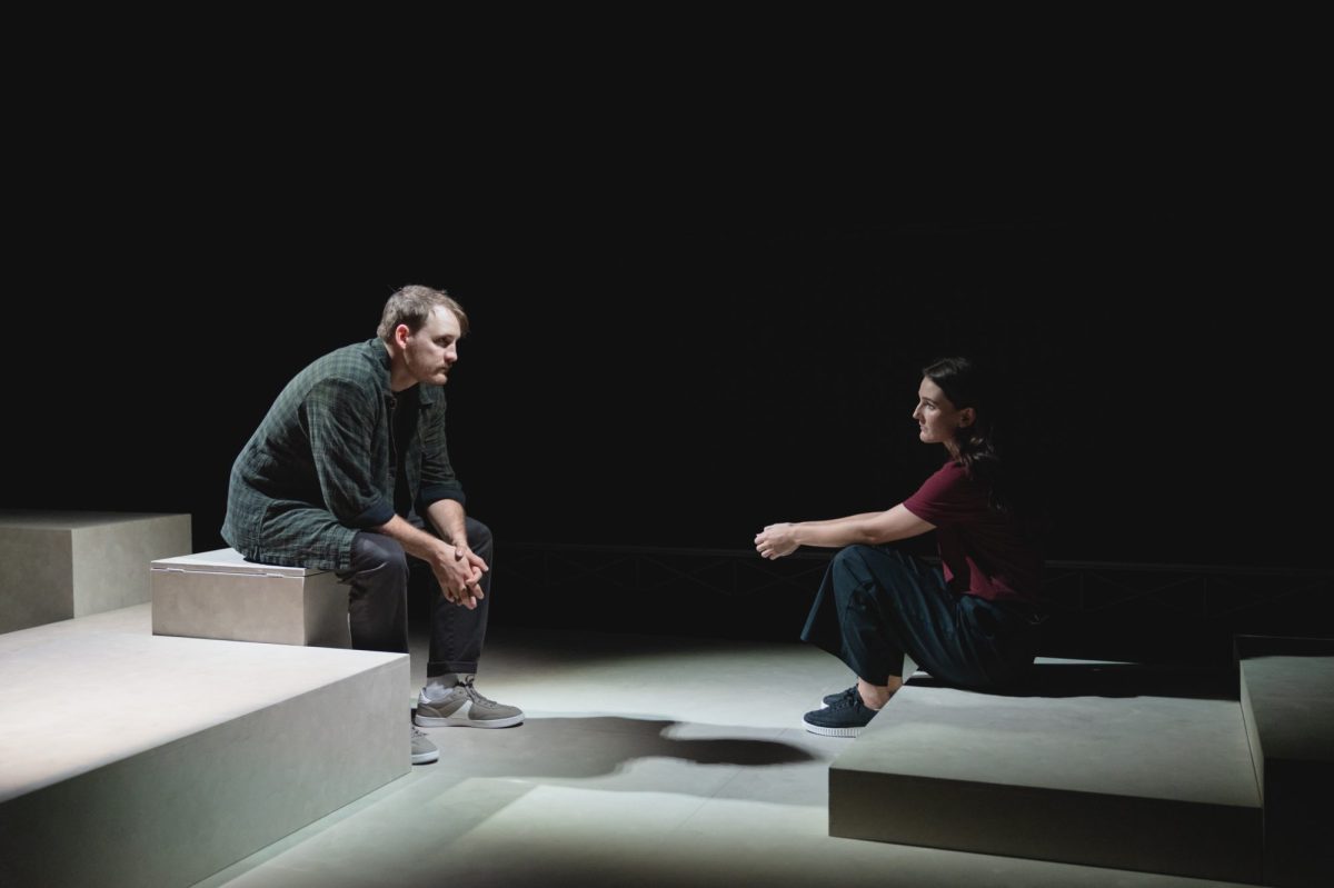 Shadow of Doubt: a young male and a female actor sitting on stage facing each other on a minimal set made of grey blocks. They sit in front of a dark backdrop.