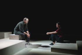 Shadow of Doubt: a young male and a female actor sitting on stage facing each other on a minimal set made of grey blocks. They sit in front of a dark backdrop.