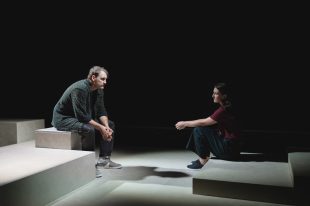 Shadow of Doubt: a young male and a female actor sitting on stage facing each other on a minimal set made of grey blocks. They sit in front of a dark backdrop.