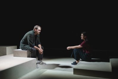 Shadow of Doubt: a young male and a female actor sitting on stage facing each other on a minimal set made of grey blocks. They sit in front of a dark backdrop.