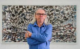 Andrew Tomkins (an older Caucasian man with long gray hair and a blue shirt) standing in front of a large artwork with rusty colours depicting a sunken vessel and leaves.