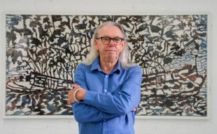 Andrew Tomkins (an older Caucasian man with long gray hair and a blue shirt) standing in front of a large artwork with rusty colours depicting a sunken vessel and leaves.