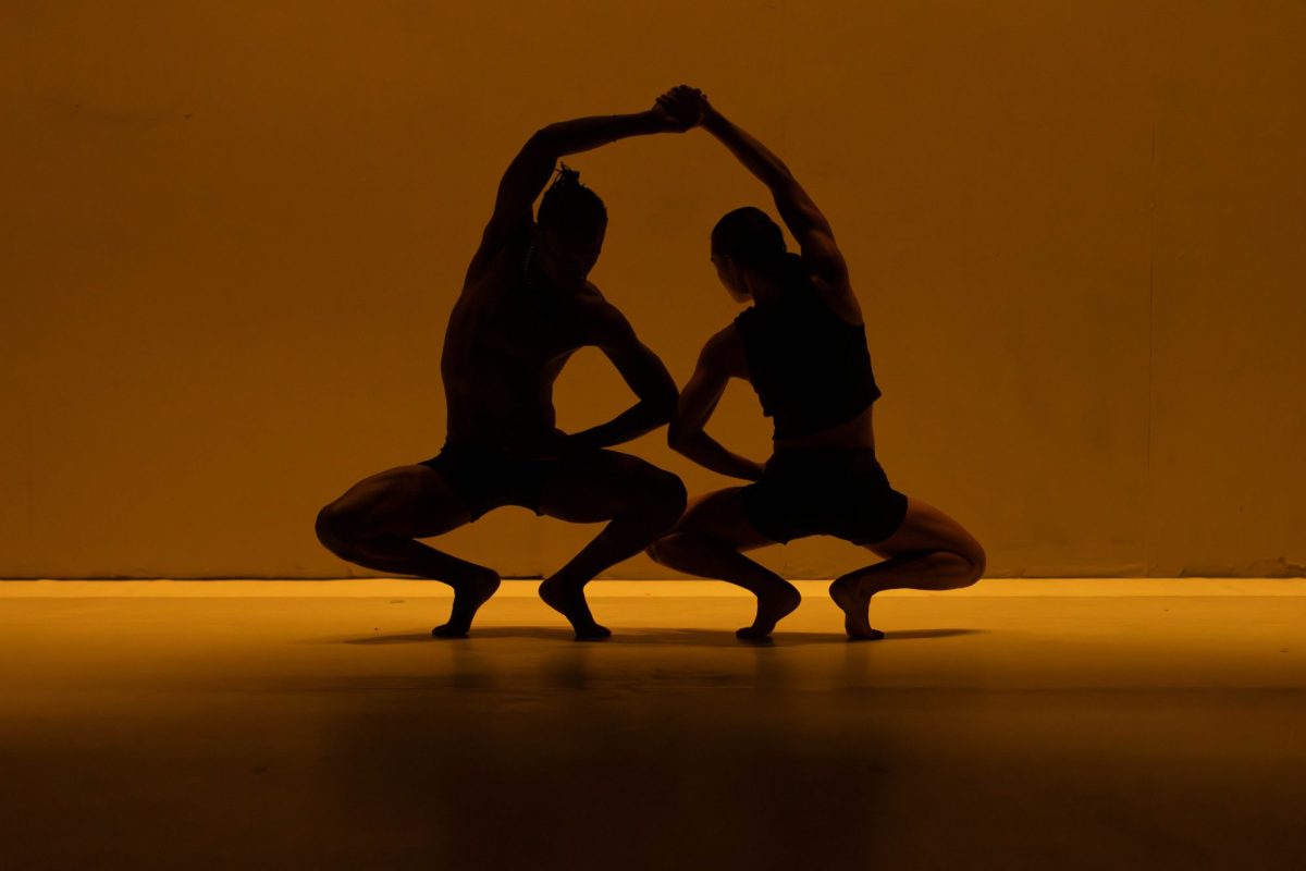 In yellow light, a silhouette of two dancers crouching, with their arms over their heads.