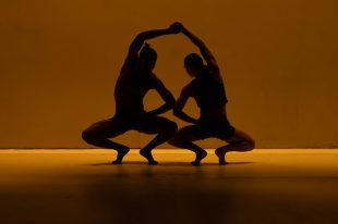 In yellow light, a silhouette of two dancers crouching, with their arms over their heads.