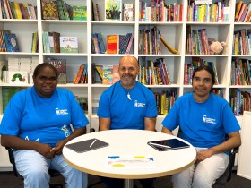 Three indigenous people in bright blue t-shirts sitting in front of shelve of books. Indigenous Literacy