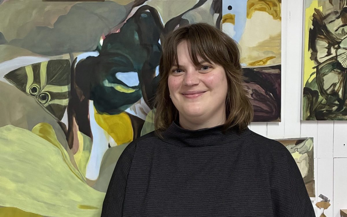 A young woman with fair skin and medium length brown hair standing in front of abstract landscape paintings and smiling.