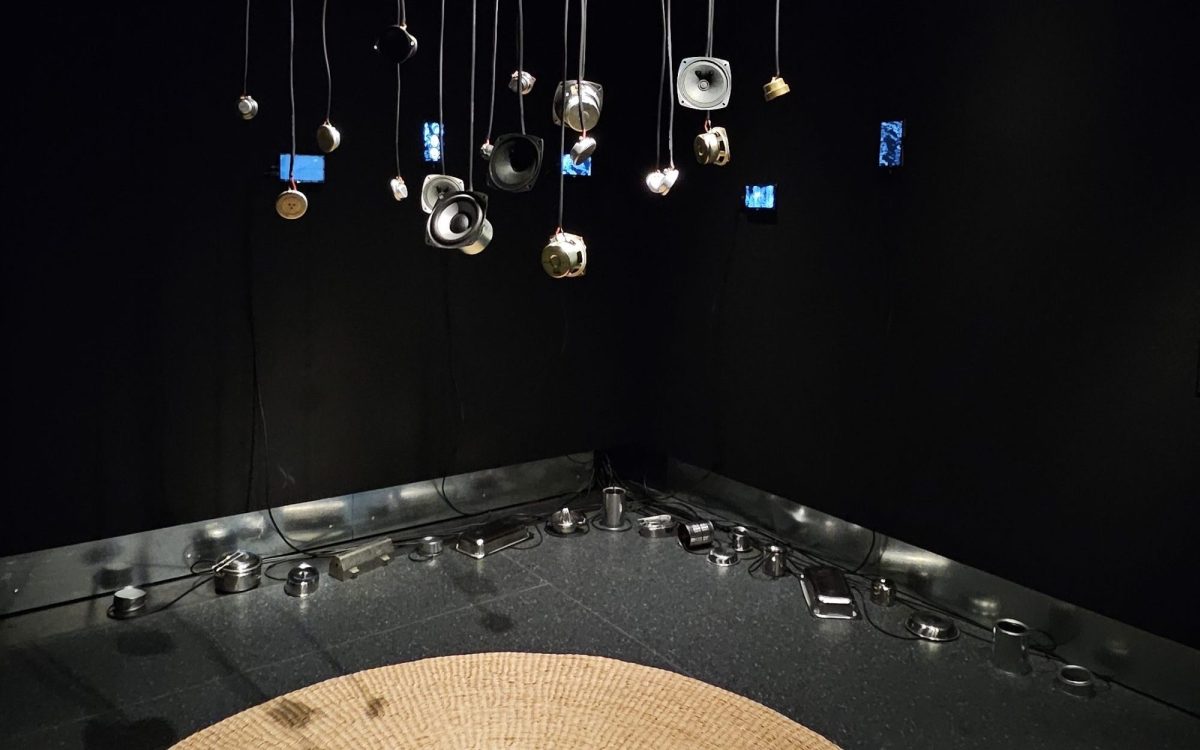 Small deconstructed speakers dangling from the ceiling above a round weaved floor mat. Aluminium containers are placed around the corner of a black wall to the back.