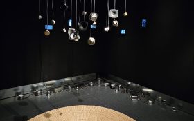 Small deconstructed speakers dangling from the ceiling above a round weaved floor mat. Aluminium containers are placed around the corner of a black wall to the back.