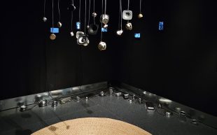 Small deconstructed speakers dangling from the ceiling above a round weaved floor mat. Aluminium containers are placed around the corner of a black wall to the back.