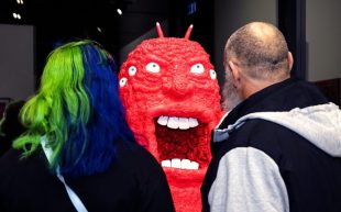 Two figures with their backs to the camera looking at a sculpture that appears to be a screaming red slug with a human face.