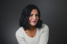 A young woman with shoulder length dark hair. She is wearing a white top.