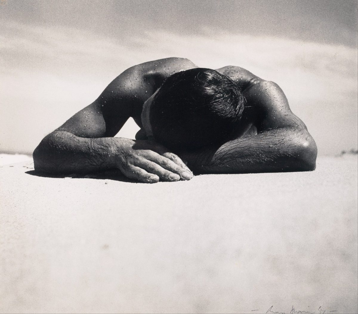 Max Dupain Sunbaker. Black and white image of a man lying in the sun, taken from ground level looking at the top of his head.