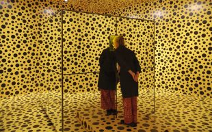 A person looking into a mirrored cube. Around the figure every surface is filled with black polka dots on a yellow background.