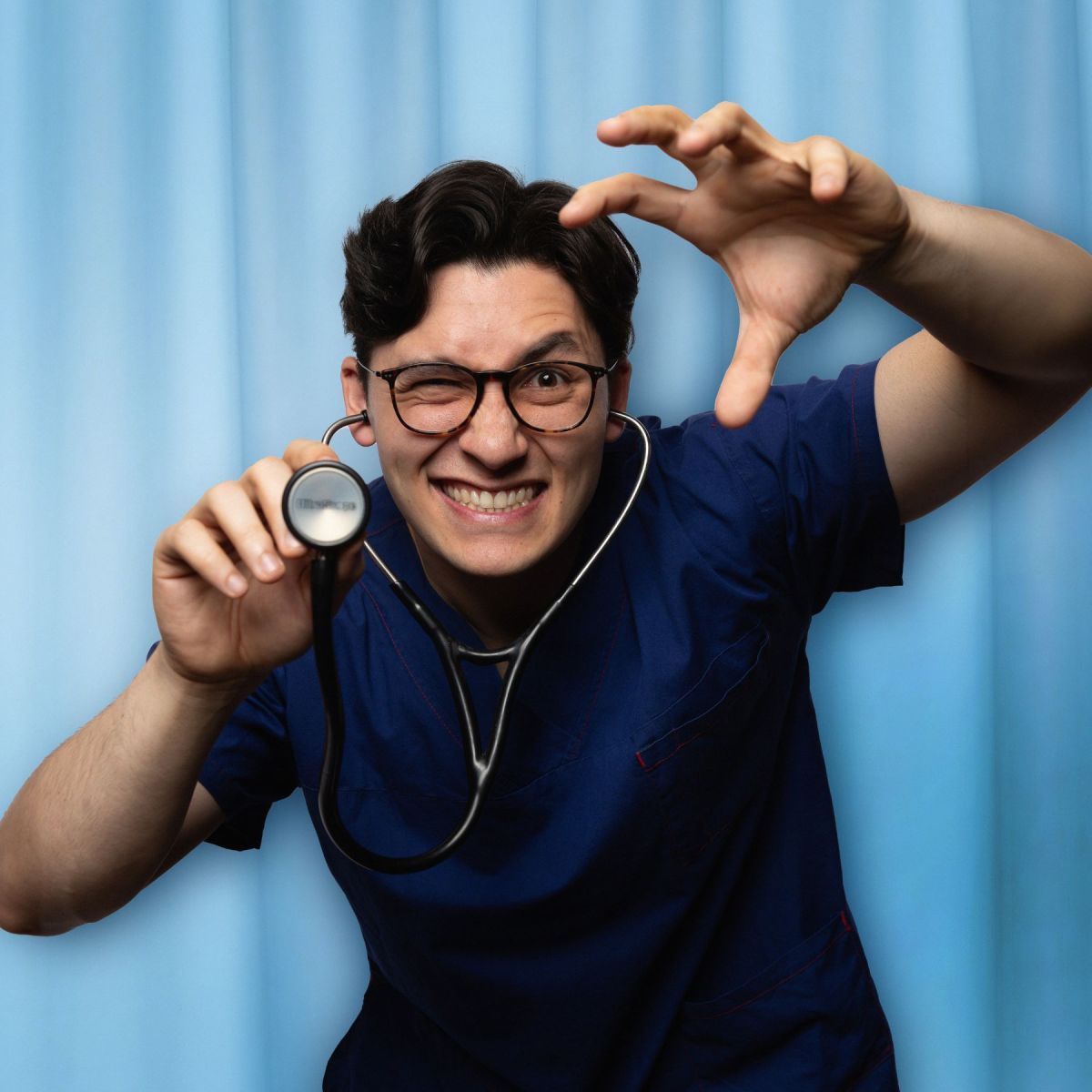 In front of a light blue curtain, a man of Asian appearance is dressed in dark blue scrubs with glasses and a stethoscope around his neck that he is brandishing at the camera, with a gleeful grimace on his face.