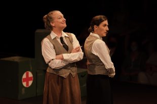 Two women wearing white shirts, olive vests and brown skirts. There is a red cross behind them in the left hand corner.