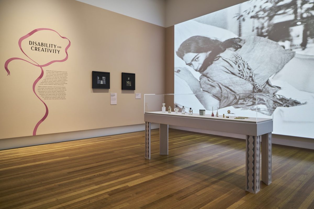 A large black and white video still is on display at the back featuring a woman, Frida Kahlo, lying on her front on a bed. In the gallery space there is also a white display table with small items on it.