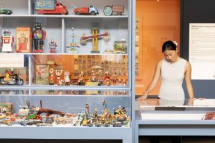 A cabinet housing small tin toys on the left. On the right is and Asian woman in white looking down at a table with a glass top.